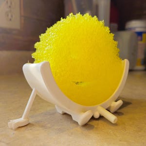 May include: A yellow kitchen sponge in a white plastic holder. The sponge is round and textured, and the holder has a curved design with a leg and two feet. The holder is designed to keep the sponge elevated.