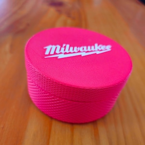 May include: A bright pink, cylindrical container with the white text "Milwaukee" on the top. The container has a textured surface and sits on a wooden surface.
