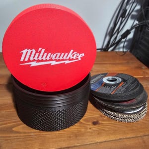 May include: A red Milwaukee branded disc with a black container and a stack of grinding discs. The red disc has the Milwaukee logo in white. The black container has a textured grip. The grinding discs are black and silver.