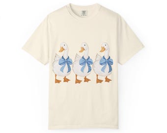 May include: Cream-colored t-shirt featuring a whimsical illustration of three white geese. Each goose is adorned with a blue and white checkered bow. The geese have orange beaks and feet. The shirt has a classic crew neck and short sleeves.