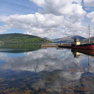 Könnte beinhalten: Ein rot-weißes Boot liegt an einem Steg mit einem Gebirge im Hintergrund. Das Wasser ist ruhig und spiegelt den Himmel und die Wolken wider.
