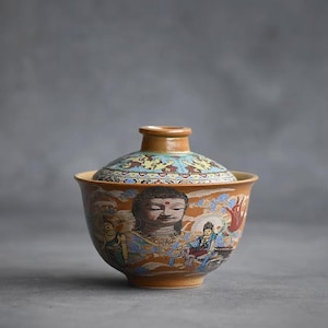 May include: A brown ceramic teacup with a lid, featuring a colorful painted design of a Buddha and other figures. The cup has a wide rim and a rounded body.
