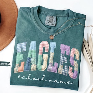 May include: Blue spruce Comfort Colors t-shirt with the word "EAGLES" in patchwork letters. The letters are in various patterns and colors. The words "school name" are written in cursive below the word "EAGLES". A gold necklace is draped across the shirt.