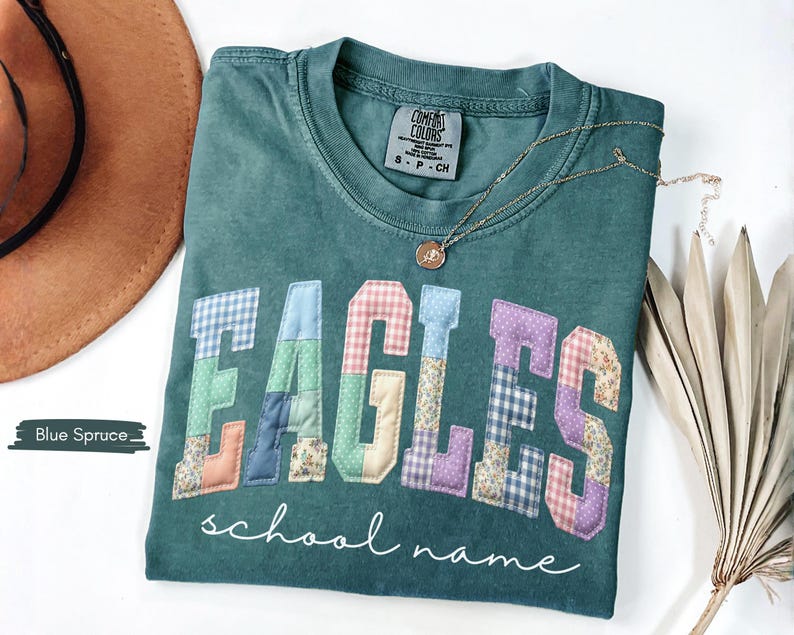 May include: Blue spruce Comfort Colors t-shirt with the word "EAGLES" in patchwork letters. The letters are in various patterns and colors. The words "school name" are written in cursive below the word "EAGLES". A gold necklace is draped across the shirt.