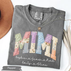 May include: A gray Comfort Colors t-shirt with the word "MIMI" in colorful patchwork letters. Below the word are names: Sophia, James, Noah, Emily, and Olivia. A silver necklace with a small pendant is on the shirt.