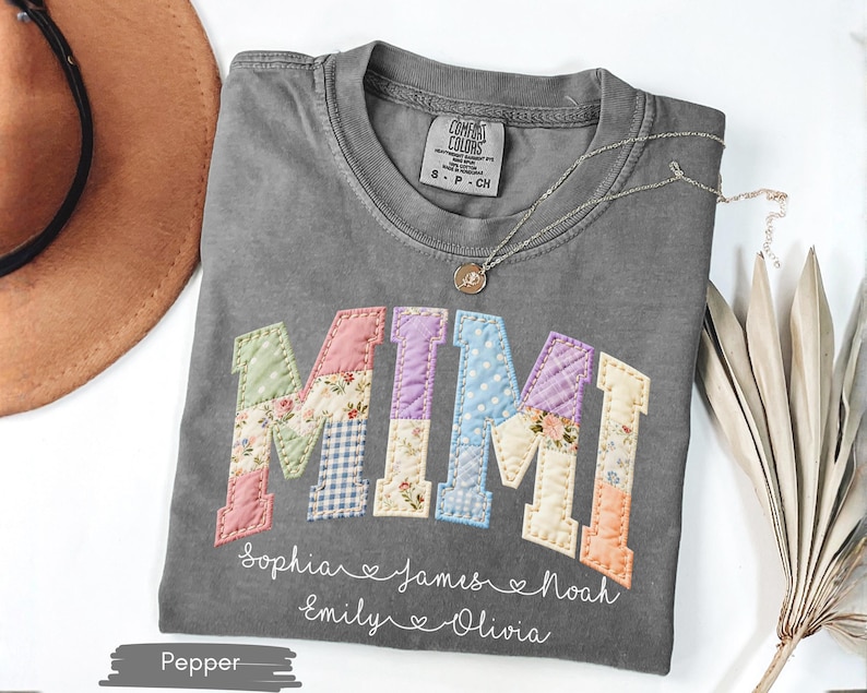 May include: A gray Comfort Colors t-shirt with the word "MIMI" in colorful patchwork letters. Below the word are names: Sophia, James, Noah, Emily, and Olivia. A silver necklace with a small pendant is on the shirt.
