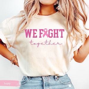 K&ouml;nnte beinhalten: Elfenbeinfarbenes T-Shirt mit den Worten "WE FIGHT" in kr&auml;ftigen rosa Buchstaben, einer rosa Schleife und dem Wort "together" in Schreibschrift. Das Shirt wird mit einer blauen Jeans getragen.