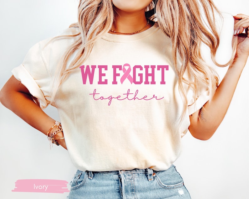 K&ouml;nnte beinhalten: Elfenbeinfarbenes T-Shirt mit den Worten "WE FIGHT" in kr&auml;ftigen rosa Buchstaben, einer rosa Schleife und dem Wort "together" in Schreibschrift. Das Shirt wird mit einer blauen Jeans getragen.