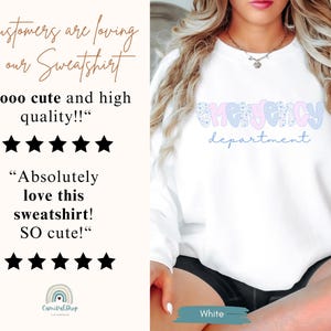 May include: White sweatshirt with the words "Emergency Department" in a playful font. The word "Emergency" is in pink and blue patterned letters, and "department" is in a light blue script. The sweatshirt is part of a clothing collection.