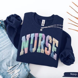 May include: Navy blue sweatshirt with the word "NURSE" in colorful patchwork letters. A silver necklace with a small pendant rests on the sweatshirt. Light blue ripped jeans are visible on the left side of the image.