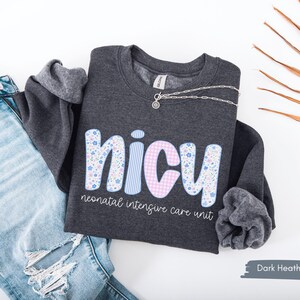 Op de afbeelding: Donkergrijs sweatshirt met het woord "NICU" in een bloemen-, gestreept- en ruitontwerp. De tekst "neonatal intensive care unit" staat onder het woord "NICU". Een zilveren ketting ligt op de trui. Lichtblauwe jeans staan op de achtergrond.