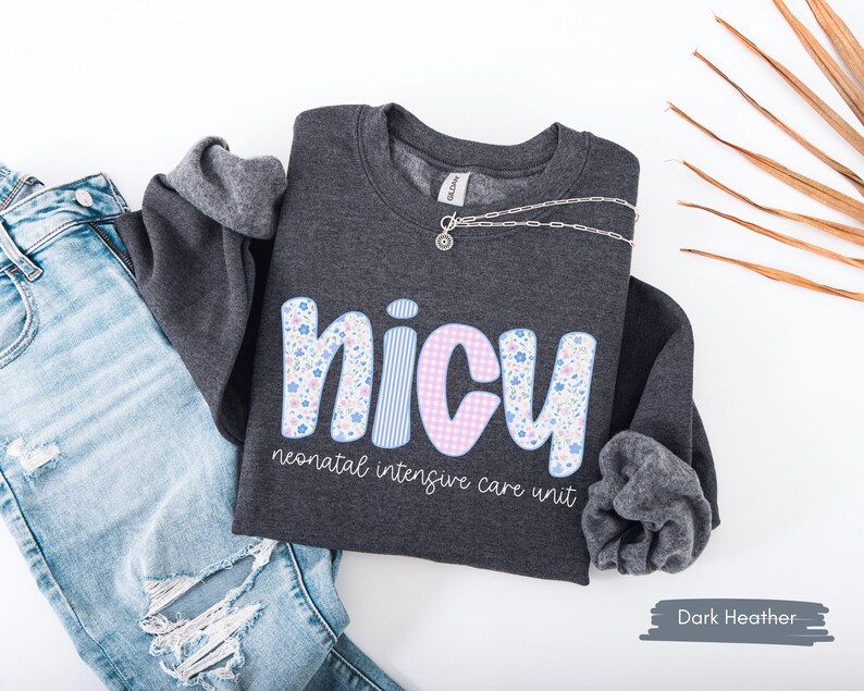 Op de afbeelding: Donkergrijs sweatshirt met het woord "NICU" in een bloemen-, gestreept- en ruitontwerp. De tekst "neonatal intensive care unit" staat onder het woord "NICU". Een zilveren ketting ligt op de trui. Lichtblauwe jeans staan op de achtergrond.