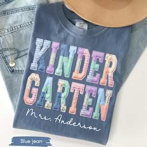 May include: A blue t-shirt with the word "KINDERGARTEN" in colorful patchwork letters. The name "Mrs. Anderson" is printed below the word. The shirt is laid on top of blue jeans and a tan hat.