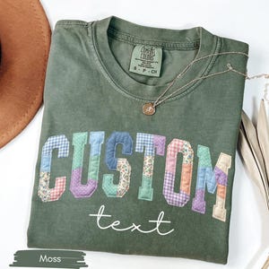 May include: A moss green t-shirt with the word "CUSTOM" in patchwork letters and the word "text" in cursive. A gold necklace and a brown hat are also visible.