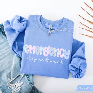 May include: A Carolina blue sweatshirt with the words "emergency department" in a playful font. The word "emergency" is in a mix of patterns and colors. A silver necklace with a small pendant rests on the shirt. Light blue jeans are visible in the background.