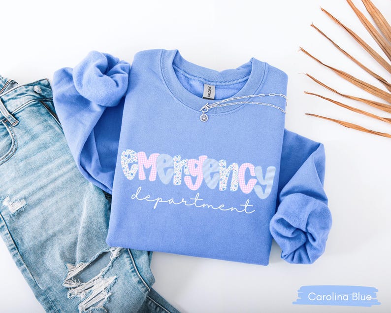 May include: A Carolina blue sweatshirt with the words "emergency department" in a playful font. The word "emergency" is in a mix of patterns and colors. A silver necklace with a small pendant rests on the shirt. Light blue jeans are visible in the background.