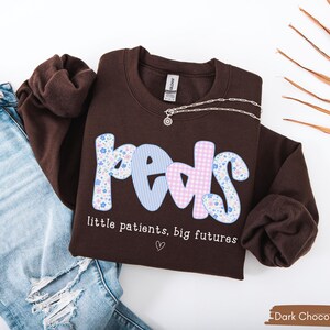 May include: A dark chocolate-colored sweatshirt with the word "Peds" in a playful font, featuring floral, striped, and gingham patterns. The text "little patients, big futures" is below the word "Peds". A silver necklace is draped on the sweatshirt.
