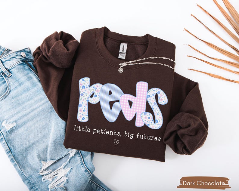 May include: A dark chocolate-colored sweatshirt with the word "Peds" in a playful font, featuring floral, striped, and gingham patterns. The text "little patients, big futures" is below the word "Peds". A silver necklace is draped on the sweatshirt.