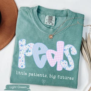 May include: Light green t-shirt with the word "peds" in a floral, gingham, and striped design. The text "little patients, big futures" is below the word "peds". A gold necklace and a brown hat are also in the image.