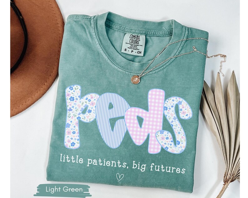 May include: Light green t-shirt with the word "peds" in a floral, gingham, and striped design. The text "little patients, big futures" is below the word "peds". A gold necklace and a brown hat are also in the image.