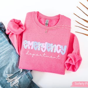May include: A pink crewneck sweatshirt with the words "emergency department" in a playful font. The word "emergency" is in a light blue and patterned design. A silver necklace and distressed blue jeans are next to the sweatshirt.