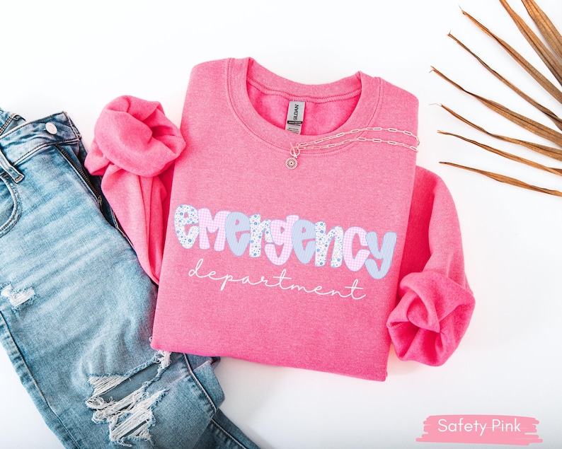 May include: A pink crewneck sweatshirt with the words "emergency department" in a playful font. The word "emergency" is in a light blue and patterned design. A silver necklace and distressed blue jeans are next to the sweatshirt.