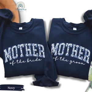 May include: Two navy blue sweatshirts with the text "MOTHER of the bride" and "MOTHER of the groom" in white, floral-patterned lettering. The sweatshirts are displayed on a white surface.