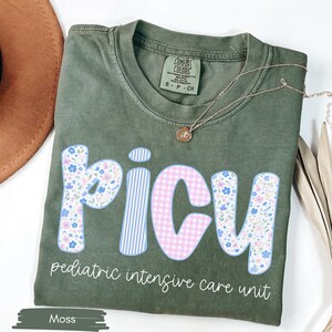 Puede incluir: Camiseta verde musgo con la palabra "PICU" en una fuente l&uacute;dica, con estampados florales, a rayas y cuadros. El texto "pediatric intensive care unit" est&aacute; debajo. Un collar dorado est&aacute; drapeado sobre la camiseta.
