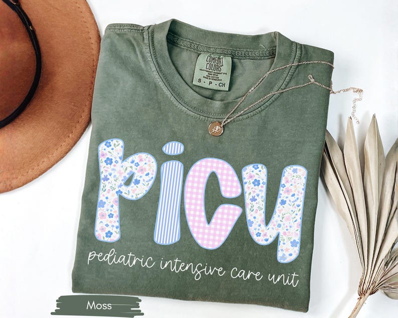 Puede incluir: Camiseta verde musgo con la palabra "PICU" en una fuente l&uacute;dica, con estampados florales, a rayas y cuadros. El texto "pediatric intensive care unit" est&aacute; debajo. Un collar dorado est&aacute; drapeado sobre la camiseta.