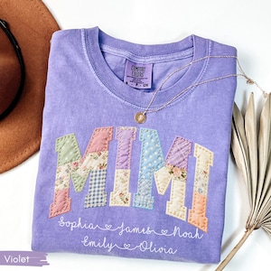 May include: A violet Comfort Colors t-shirt with the word "MIMI" in patchwork letters. The letters are in pastel colors and floral patterns. Below the word are names. A gold necklace and a brown hat are in the background.