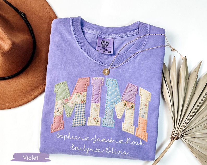 May include: A violet Comfort Colors t-shirt with the word "MIMI" in patchwork letters. The letters are in pastel colors and floral patterns. Below the word are names. A gold necklace and a brown hat are in the background.