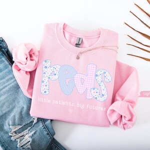 May include: A light pink sweatshirt with the word "PEDS" in a floral and gingham pattern. The text "little patients, big futures" is below the word. The sweatshirt is next to a pair of blue jeans.