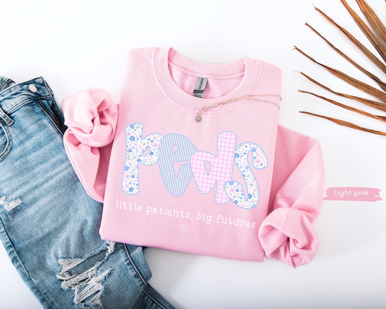 May include: A light pink sweatshirt with the word "PEDS" in a floral and gingham pattern. The text "little patients, big futures" is below the word. The sweatshirt is next to a pair of blue jeans.