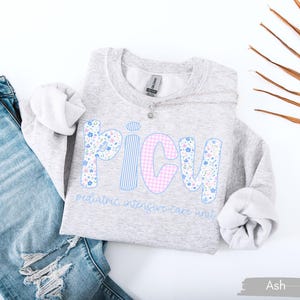 May include: A light gray sweatshirt with the word "PICU" in a floral, striped, and gingham design. The text "pediatric intensive care unit" is below the word. The sweatshirt is paired with blue jeans.