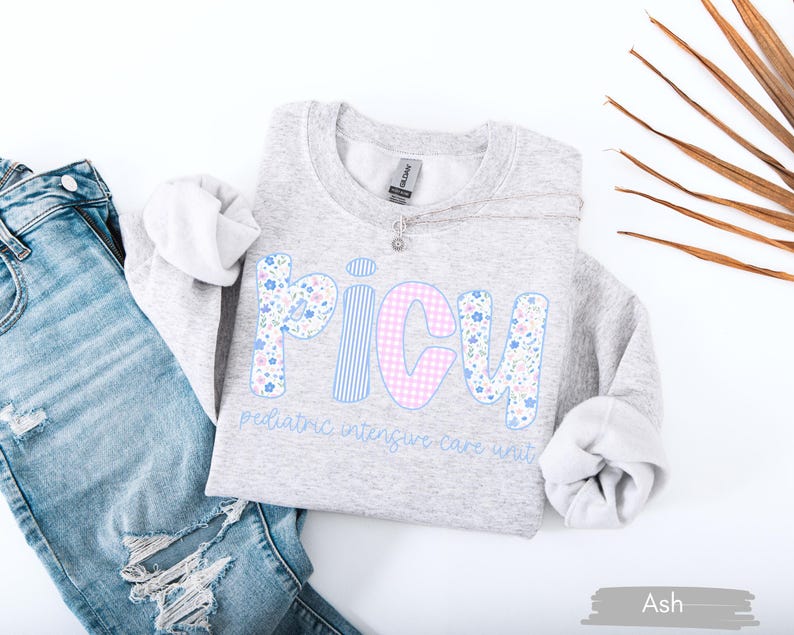 May include: A light gray sweatshirt with the word "PICU" in a floral, striped, and gingham design. The text "pediatric intensive care unit" is below the word. The sweatshirt is paired with blue jeans.