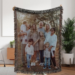 May include: A woven throw blanket featuring a family portrait. The image shows a group of people in a natural outdoor setting. The blanket has a dark fringe and is being held up, showcasing the full image.