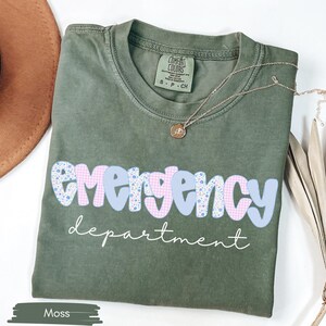 May include: Moss green t-shirt with the words "emergency department" in a playful font. The word "emergency" is in a blue and pink floral and gingham pattern. A gold necklace rests on the shirt.