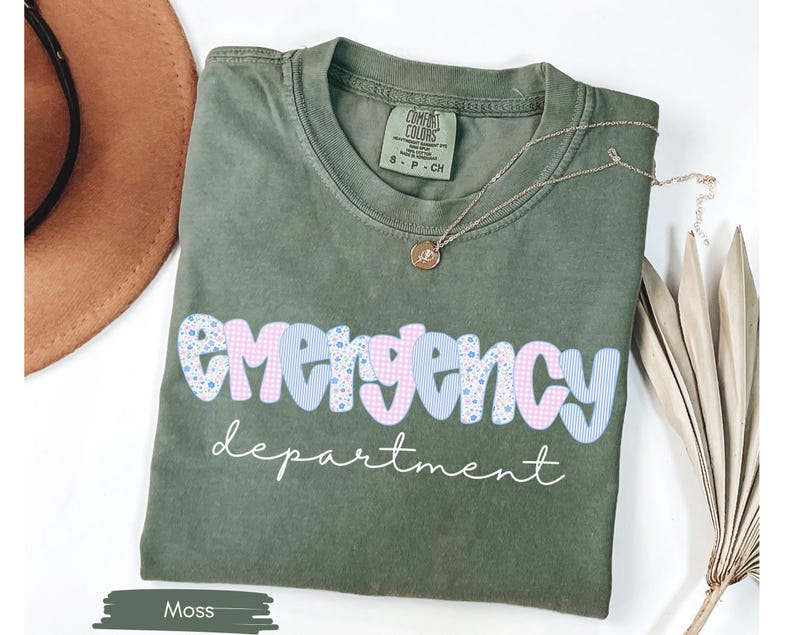May include: Moss green t-shirt with the words "emergency department" in a playful font. The word "emergency" is in a blue and pink floral and gingham pattern. A gold necklace rests on the shirt.