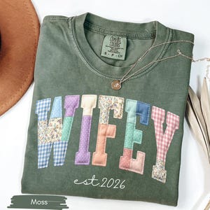 May include: A moss green t-shirt with the word "WIFEY" in patchwork letters. The letters are made of various patterned fabrics, including floral and gingham. The text "est. 2026" is below the word "WIFEY". A gold necklace is draped on the shirt.