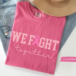 K&ouml;nnte beinhalten: Ein pinkfarbenes T-Shirt mit den Worten "WE FIGHT together" in hellrosa Schrift und einer rosa Schleifengrafik. Das Shirt liegt gefaltet auf einer Jeans und einem beigen Hut.