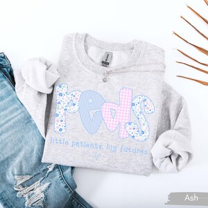 May include: A light gray sweatshirt with the word "PEDS" in blue, pink, and floral patterns. The text "little patients, big futures" is below the word. A pair of blue jeans is next to the sweatshirt.