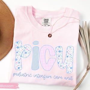 Puede incluir: Camiseta rosa claro con la palabra "PICU" en patrones florales, a rayas y cuadros. El texto "pediatric intensive care unit" est&aacute; debajo. Tambi&eacute;n se ven un collar dorado y un sombrero marr&oacute;n.