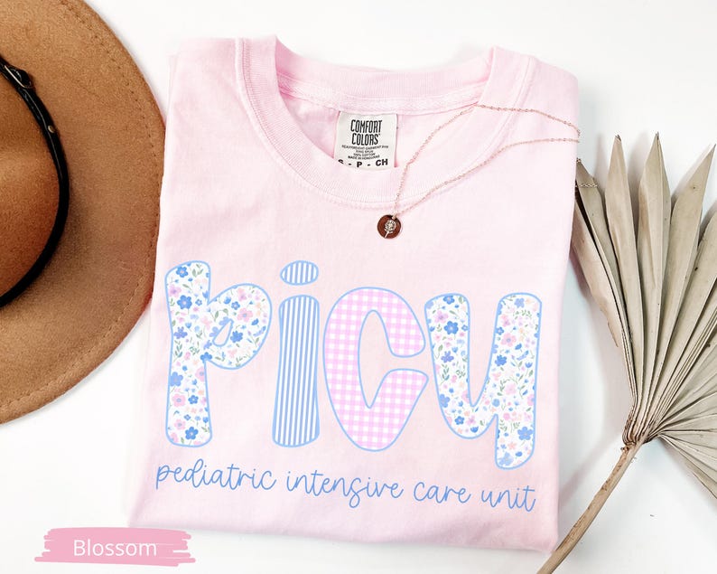 Puede incluir: Camiseta rosa claro con la palabra "PICU" en patrones florales, a rayas y cuadros. El texto "pediatric intensive care unit" est&aacute; debajo. Tambi&eacute;n se ven un collar dorado y un sombrero marr&oacute;n.