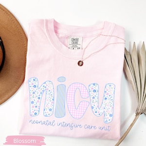 Peut inclure: Un t-shirt rose clair avec le mot "NICU" dans une police fantaisiste. Les lettres sont d&eacute;cor&eacute;es de motifs floraux, ray&eacute;s et vichy. Le texte "neonatal intensive care unit" est en dessous. Un collier dor&eacute; et un chapeau sont &eacute;galement dans l'image.