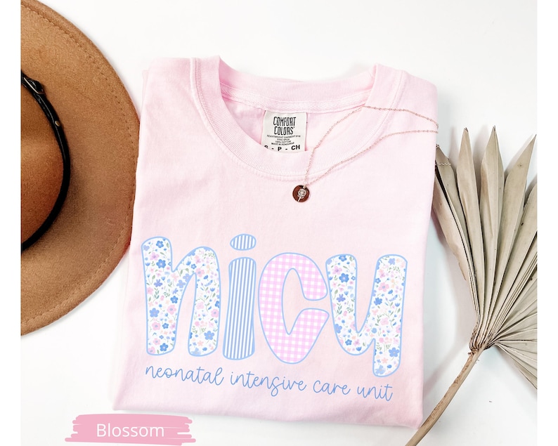 Peut inclure: Un t-shirt rose clair avec le mot "NICU" dans une police fantaisiste. Les lettres sont d&eacute;cor&eacute;es de motifs floraux, ray&eacute;s et vichy. Le texte "neonatal intensive care unit" est en dessous. Un collier dor&eacute; et un chapeau sont &eacute;galement dans l'image.