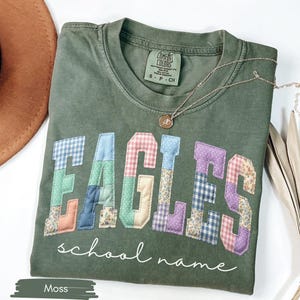 May include: A moss green t-shirt with the word "EAGLES" in patchwork letters. The letters are made of various patterned fabrics. Below the word "EAGLES" is the phrase "school name" in a cursive font. A gold necklace is draped across the shirt.