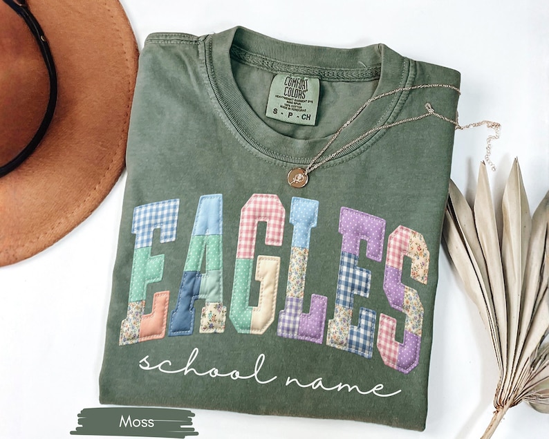 May include: A moss green t-shirt with the word "EAGLES" in patchwork letters. The letters are made of various patterned fabrics. Below the word "EAGLES" is the phrase "school name" in a cursive font. A gold necklace is draped across the shirt.