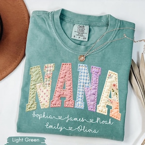 May include: Light green t-shirt with the word "NANA" in patchwork letters. The letters are in various floral and patterned fabrics. Below the word "NANA" are names: Sophia, James, Noah, Emily, and Olivia. A gold necklace rests on the shirt.