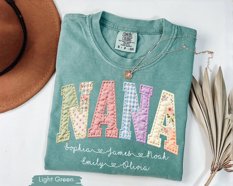 May include: Light green t-shirt with the word "NANA" in patchwork letters. The letters are in various floral and patterned fabrics. Below the word "NANA" are names: Sophia, James, Noah, Emily, and Olivia. A gold necklace rests on the shirt.