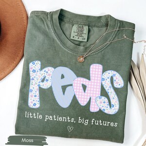 May include: Moss green t-shirt with the word "peds" in a playful font, featuring floral, striped, and gingham patterns. The text "little patients, big futures" is below the word "peds". A gold necklace and a brown hat are also in the image.